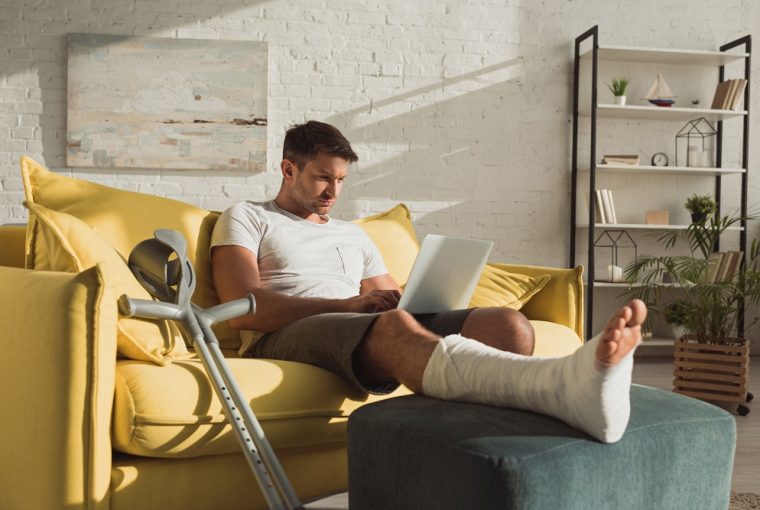 homem com perna engessada sentado no sofá mexendo no computador para solicitar o seguro DPVAT