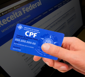 computador ao fundo mostrando o site da Receita Federal e a frente uma mão segurando um cartão de CPF, simbolizando como descobrir o CPF pelo nome