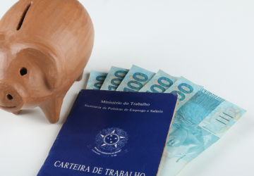 carteira de trabalho brasileira com notas de cem reais dentro e ao lado um cofre em formato de porco de madeira, simbolizando o investimento em um plano de previdência privada para a aposentadoria