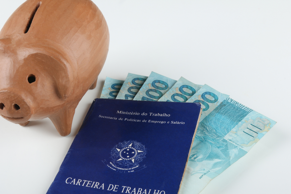 carteira de trabalho brasileira com notas de cem reais dentro e ao lado um cofre em formato de porco de madeira, simbolizando o investimento em um plano de previdência privada para a aposentadoria