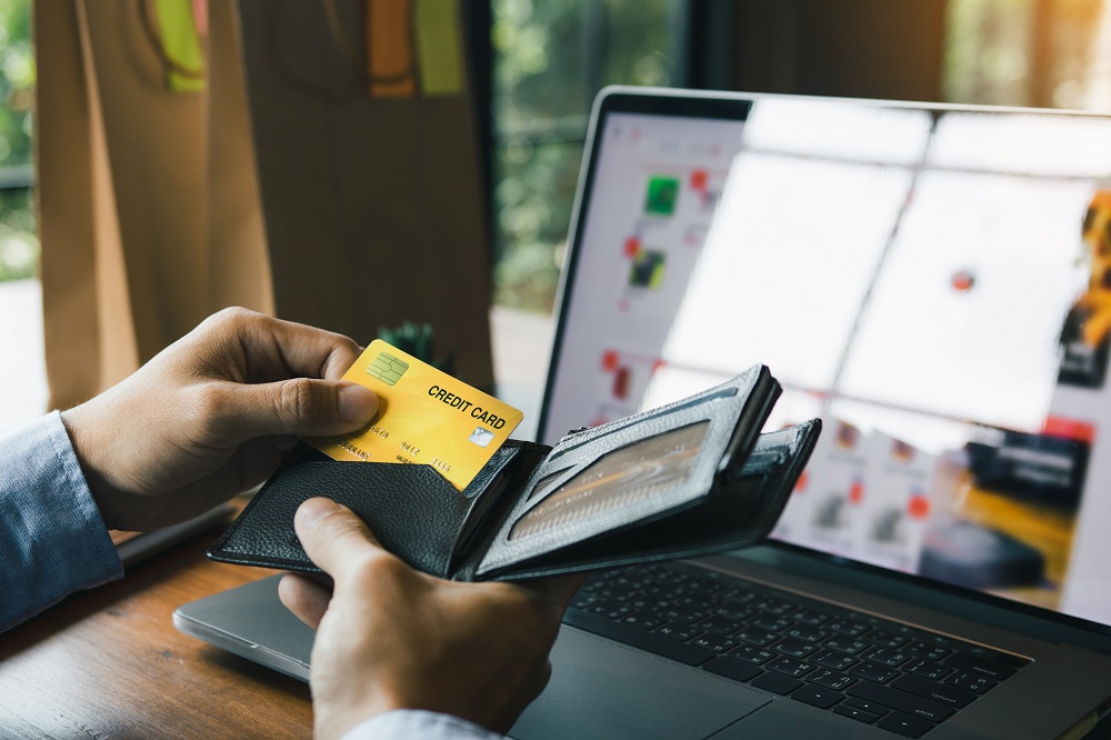 mão masculina abrindo uma carteira e tirando um cartão de crédito de dentro, em frente à um computador com compras online no site, simbolizando o estorno e cancelamento de compras no cartão