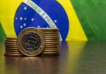 bandeira do Brasil ao fundo e em cima de uma mesa algumas moedas de um real empilhadas, simbolizando o investimento no Tesouro Direto com Aumento da Taxa Selic