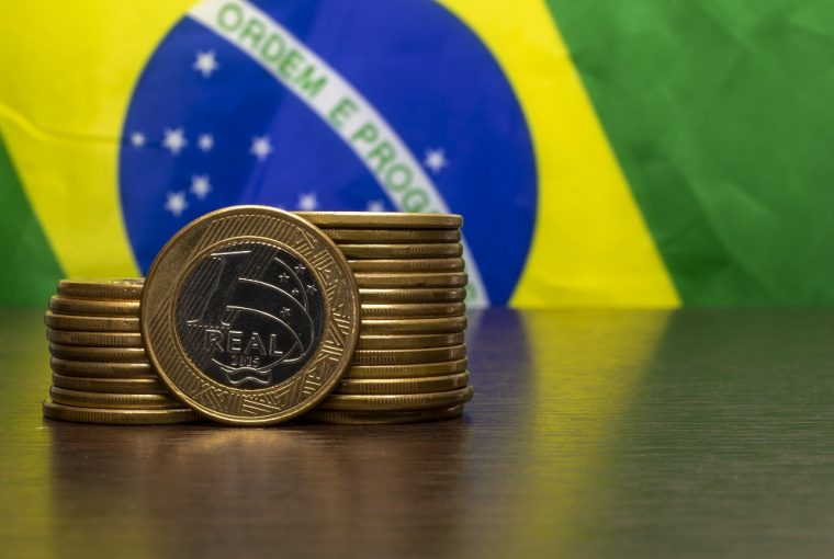 bandeira do Brasil ao fundo e em cima de uma mesa algumas moedas de um real empilhadas, simbolizando o investimento no Tesouro Direto com Aumento da Taxa Selic