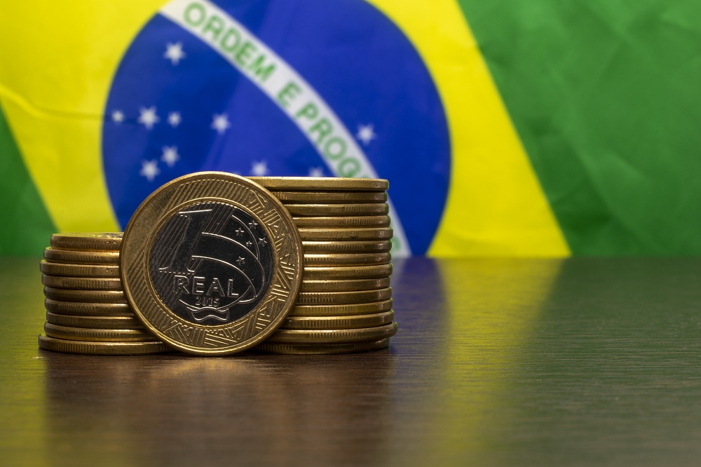 bandeira do Brasil ao fundo e em cima de uma mesa algumas moedas de um real empilhadas, simbolizando o investimento no Tesouro Direto com Aumento da Taxa Selic