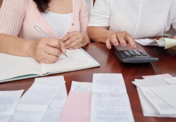 Mulheres irreconhecíveis, sentado à mesa com recibos, contando com calculadora e escrevendo no diário simulando se vale a pena parcelar contas no boleto