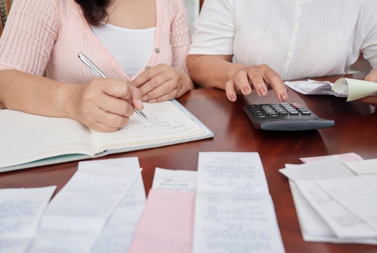 Mulheres irreconhecíveis, sentado à mesa com recibos, contando com calculadora e escrevendo no diário simulando se vale a pena parcelar contas no boleto