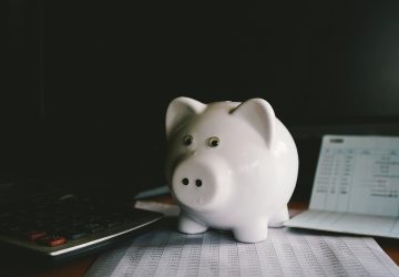 cofre de porco de porcelana branco em cima de um relatório financeiro ao lado de uma calculadora simbolizando como escolher um banco para abrir sua conta poupança
