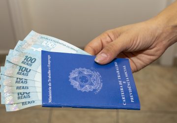 mão feminina segurando uma carteira de trabalho do Brasil com várias notas de cem reais dentro indicando o pagamento do décimo terceiro salário e como usar