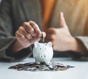 empresária fazendo sinal de mão com o polegar enquanto coloca moedas no cofrinho com uma pilha de moedas na mesa para economizar dinheiro em 2022