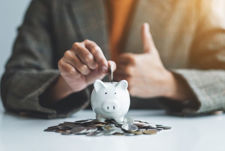 empresária fazendo sinal de mão com o polegar enquanto coloca moedas no cofrinho com uma pilha de moedas na mesa para economizar dinheiro em 2022