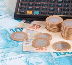 notas de cem e cinquenta reais espalhas na mesa, em cima tem moedas de um real e ao lado uma calculadora simulando qual o banco tem a menor taxa de juros para empréstimo