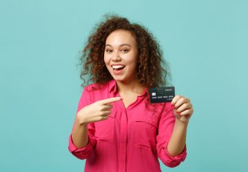 Animada garota africana em roupas casuais rosa aponta o dedo indicador no cartão do banco de crédito, simbolizando a descoberta das formas de ganhar dinheiro com o cartão de crédito