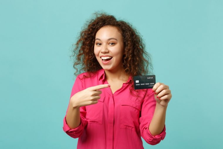 Animada garota africana em roupas casuais rosa aponta o dedo indicador no cartão do banco de crédito, simbolizando a descoberta das formas de ganhar dinheiro com o cartão de crédito