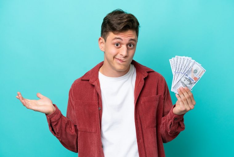 Jovem brasileiro tomando muito dinheiro isolado em fundo azul com dúvidas ao levantar as mãos simbolizando a possibilidade de Negativado pode fazer o saque-aniversário do FGTS antecipado