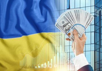 Dinheiro na mão de um homem no contexto da bandeira da ucrânia, simbolizando como o conflito e crise na Ucrânia e Rússia irá afetar a economia brasileira e mundial