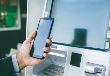 mão de homem com celular na mão na frente de um caixa eletrônico simulando o Passo a passo de como fazer o saque do FGTS pelo Pix