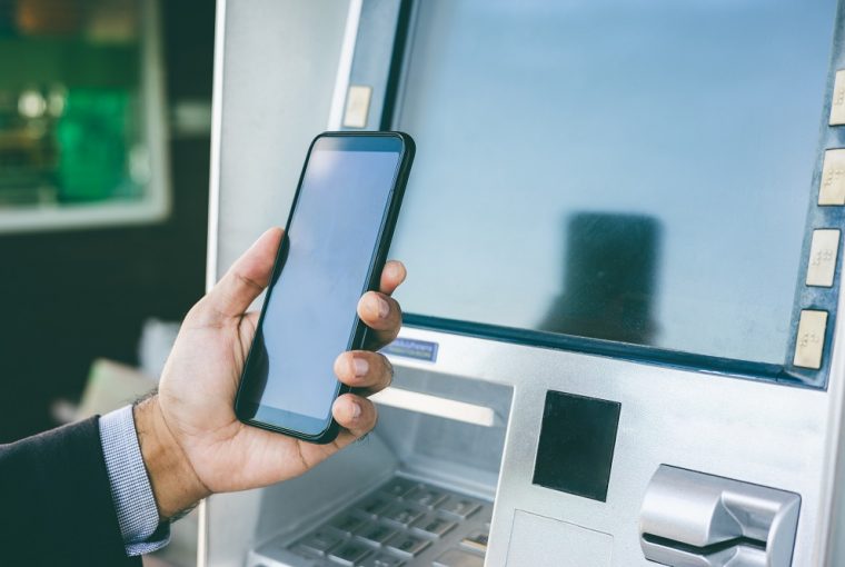 mão de homem com celular na mão na frente de um caixa eletrônico simulando o Passo a passo de como fazer o saque do FGTS pelo Pix