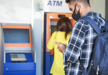 homem de máscara olhando o celular em frente à fila em um caixa ATM, simulando como fazer um saque sem cartão no caixa eletrônico