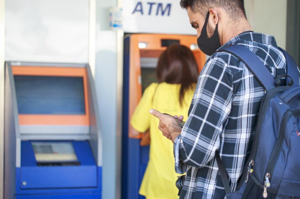 homem de máscara olhando o celular em frente à fila em um caixa ATM, simulando como fazer um saque sem cartão no caixa eletrônico