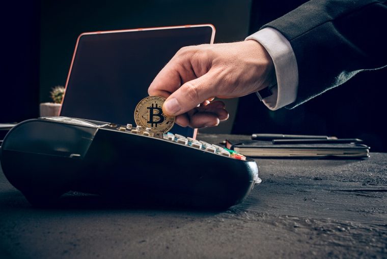maquininha de cartão onde homem está passando uma moeda de Bitcoin, simulando um pagamento, para simbolizar Onde comprar com Bitcoin no Brasil