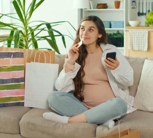 Jovem grávida faz compras on-line usando seu telefone e cartão de crédito em uma sala de estar, simbolizando a escolha do Cartão de crédito para as compras do dia das mães