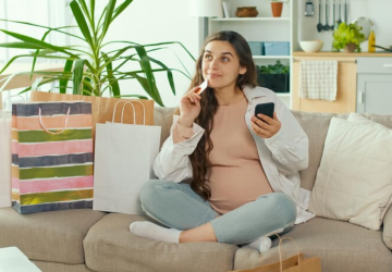 Jovem grávida faz compras on-line usando seu telefone e cartão de crédito em uma sala de estar, simbolizando a escolha do Cartão de crédito para as compras do dia das mães