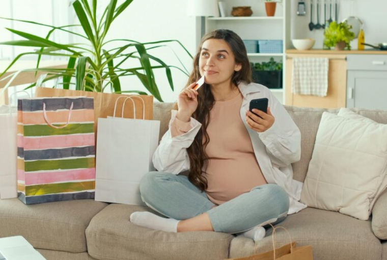 Jovem grávida faz compras on-line usando seu telefone e cartão de crédito em uma sala de estar, simbolizando a escolha do Cartão de crédito para as compras do dia das mães