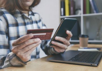 Jovem mulher segurando um cartão de crédito e usando telefone simulando como usar o seguro proteção de preço do cartão de crédito