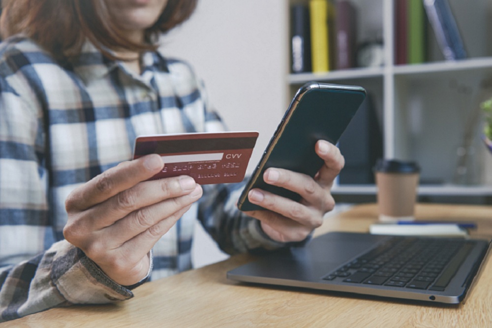 Jovem mulher segurando um cartão de crédito e usando telefone simulando como usar o seguro proteção de preço do cartão de crédito