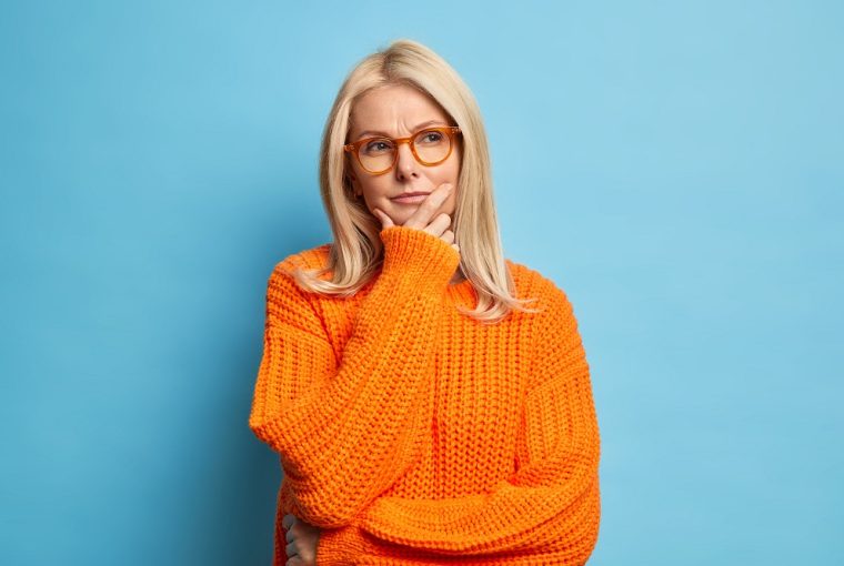 Mulher loira elegante e enrugada pensa profundamente em algo segurando o queixo, usa óculos e suéter laranja, pensando sobre se o banco pode recusar a abertura de conta
